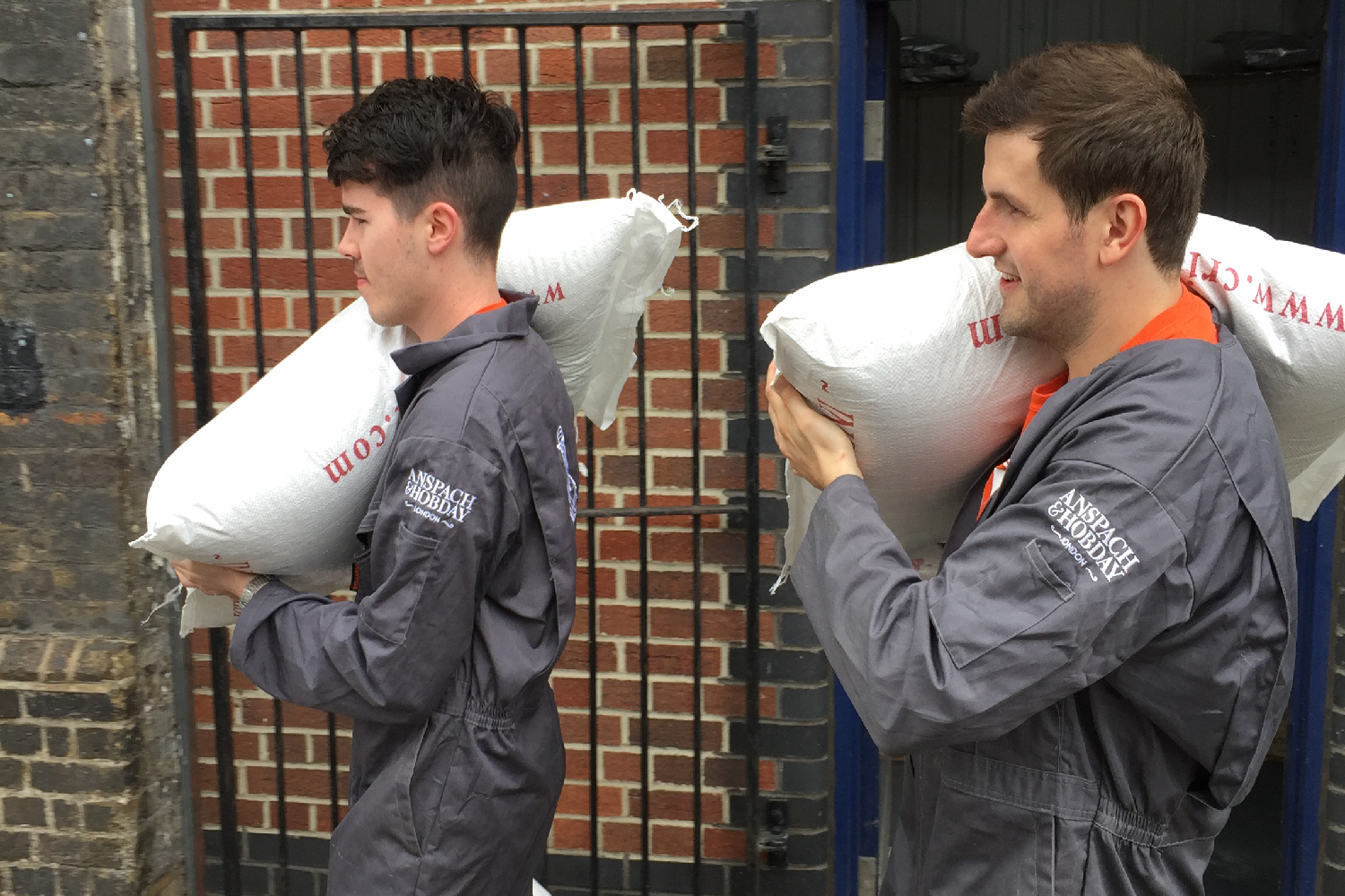 Two men in grey jumpsuits carrying sacks of grain against a brick wall background