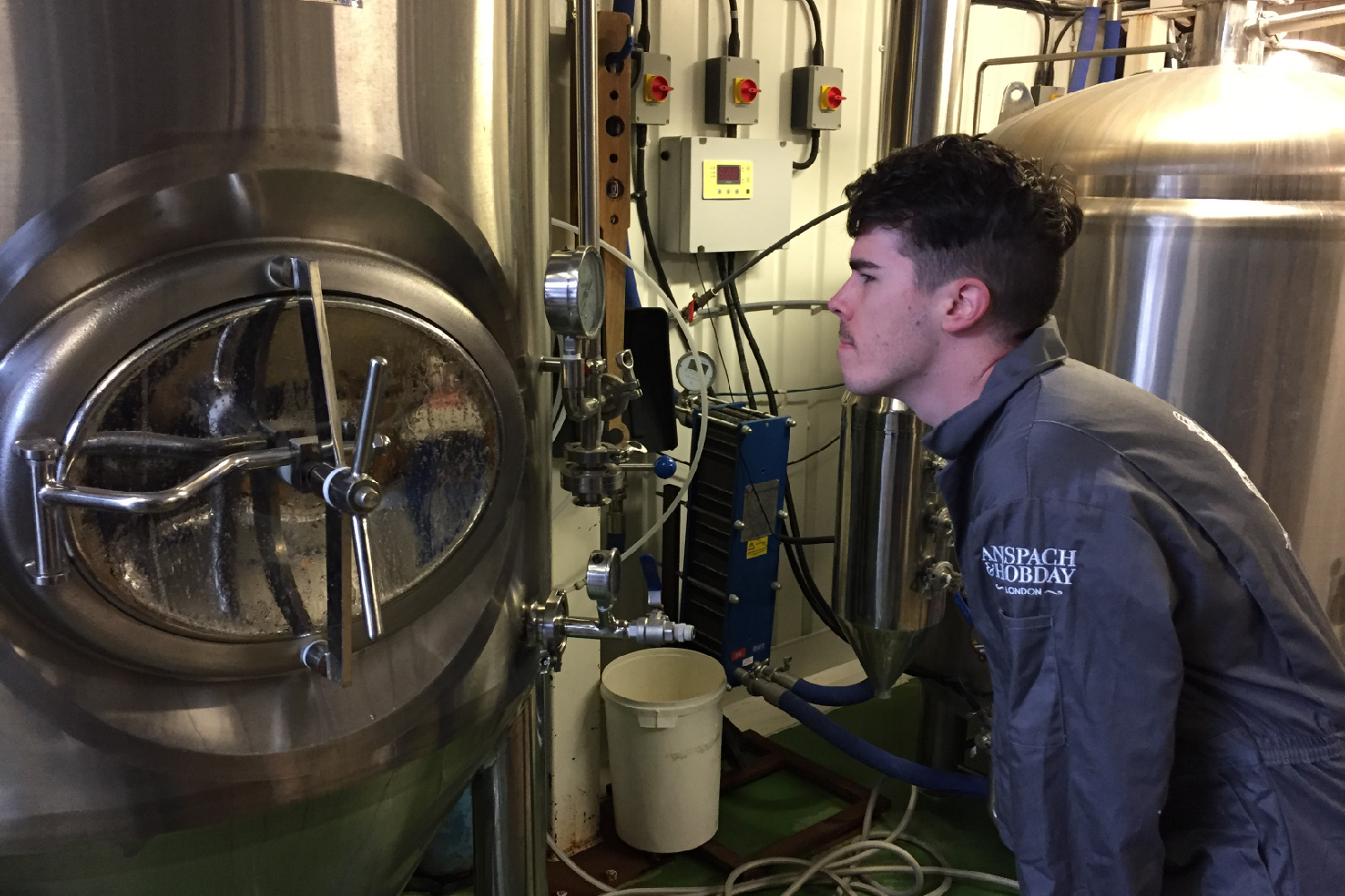 A man in a grey jumpsuit peering at a dial on a tank containing fermenting beer