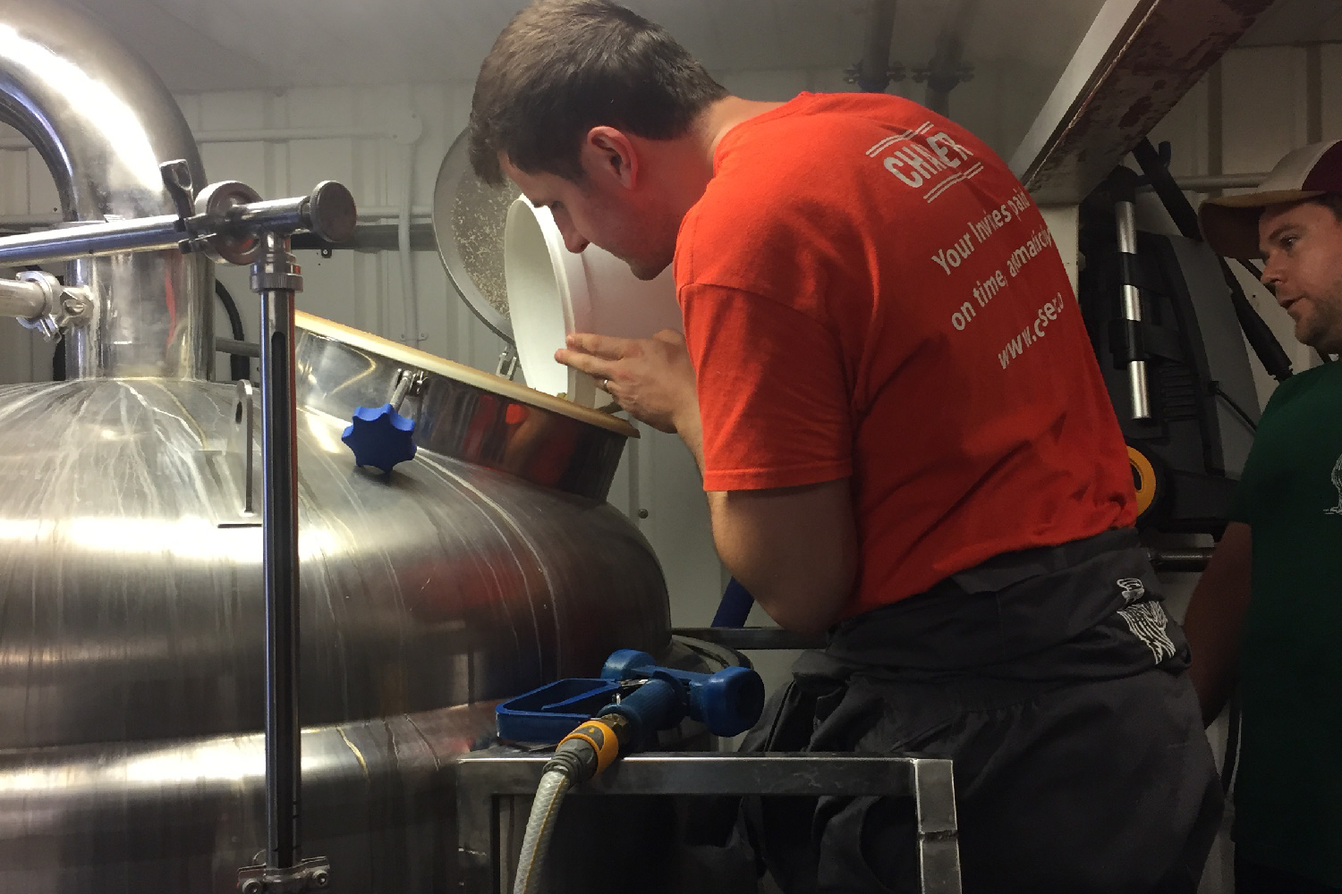 An orange-shirted man pouring hops pellets into the kettle during a beer brew