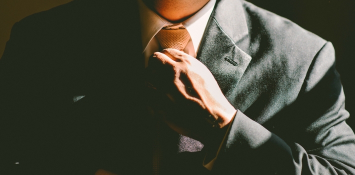 A man in a charcoal suit, bathed in shadows, adjusting his tie