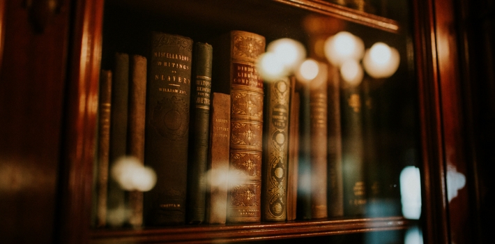 Old books on a nicely polished dark wood shelf, behind a glass cabinet door